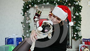 Woman hug funny pug dog in christmas costume wears Santa hat, gifts in the background