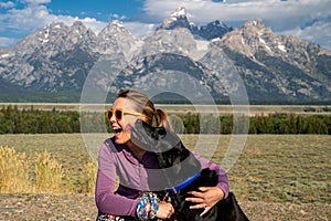 Woman hugs and holds her black labrador retriever dog in front of the Grand Teton National Park mountains in Jackson Wyoming as