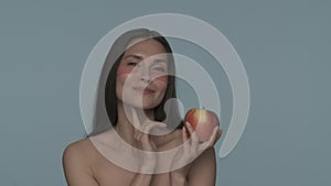 A woman with hydrogel red patches under her eyes tosses a ripe apple from one hand to the other. Seminude brunette woman