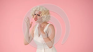 A woman in the image of Marilyn Monroe swearing in a studio against a pink background in close up.