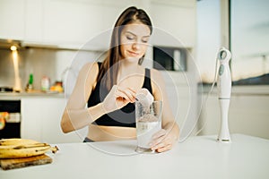 Woman with immersion blender making banana chocolate protein powder milkshake smoothie.Adding a scoop of low carb whey protein mix
