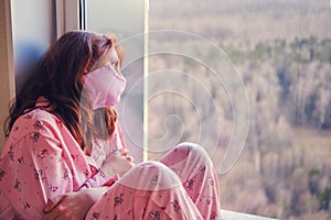 Woman infected with coronavirus wears a protective mask and standing next to the window. Pandemic caused by a flu virus, concept