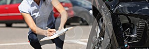 Woman inspector writing in documents on clipboard near broken car. Auto insurance