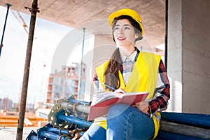 Girl in a construction helmet and vest writes in a notebook at a construction site