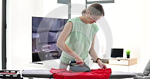 Woman ironing red shirt clothes on ironing board in laundry at home