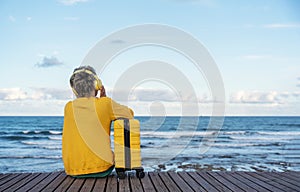 A woman in jacket with a yellow suitcase and headphones on the seaside on hot sunny day arrived in a tourist town. Take