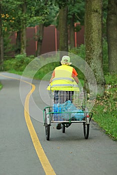 Woman janitor in overall riding bicycle with a trolley and picking up garbage in the city park