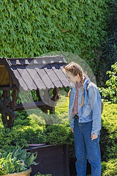 A woman in jeans clothes looks into a wooden well overgrown with green vegetation against the background of a wall with