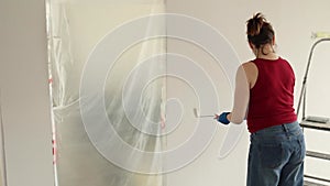 A woman in jeans dips a paint roller and paints the walls in a room white.