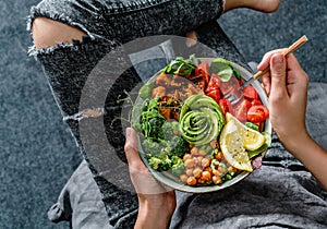 Woman in jeans holding Buddha bowl with salad, baked sweet potatoes, chickpeas, broccoli, greens, avocado, sprouts in hands.