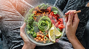 Woman in jeans holding Buddha bowl with salad, baked sweet potatoes, chickpeas, broccoli, greens, avocado, sprouts in hands.