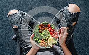 Woman in jeans holding Buddha bowl with salad, baked sweet potatoes, chickpeas, broccoli, greens, avocado, sprouts in hands.