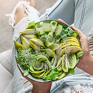 Woman in jeans holding fresh healthy greeen salad with avocado, kiwi, apple, cucumber, pear, greens and sesame on light background