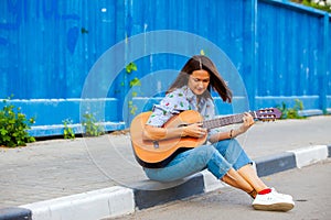 Woman in jeans sits on a road curb and plays the guitar