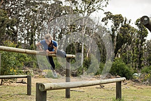 Woman vaulting over wooden beam in clearing on obstacle course wearing blue top, black leggings