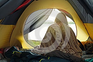 Woman just woken up in camping tent, wrapped in wool blanket and admiring sunrise on the river. Local travel on nature, trekking,