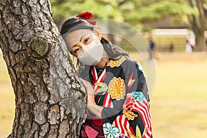 Woman in kimono wearing a mask leaned against the trunk of a pine tree.