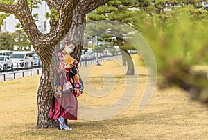 Woman in kimono wearing a mask reading a Tokyo guide book leaned against a pine tree.