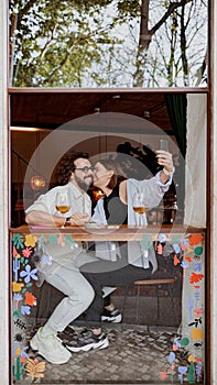 Woman kissing man in a cheek and take a selfie while they drinking wine in bar during romantic date
