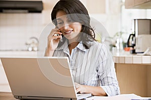 Woman in kitchen with laptop using cellular phone