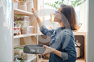 Woman in kitchen pantry. Storage wooden stand with kitchenware, products necessary to cook