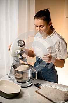Woman at kitchen prepares dough using modern electric kitchen stand mixer.