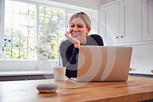 Woman In Kitchen Using Digital Smart Speaker Whilst Working From Home With Laptop During Pandemic