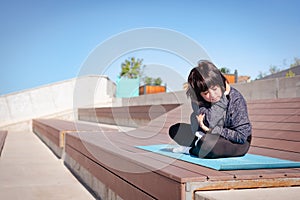 A woman kneads her body while performing yoga asanas while sitting on a wooden bench in a city park