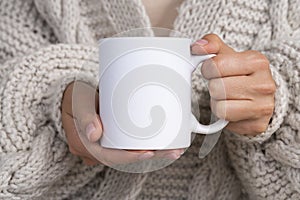 Woman in knitted sweater holding white mug in hands. Winter cup Mockup for Christmas individual gifts design