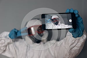 Woman with lab coat, nitrile gloves, safety glasses, face mask and NBC protection suit, taking a selfie with a smartphone and