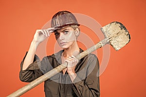 woman laborer in protective helmet and boilersuit hold shovel on orange background, construction