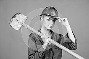 woman laborer in protective helmet and boilersuit hold shovel on orange background, construction