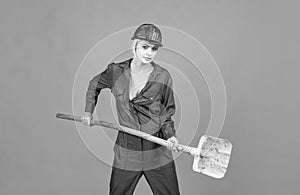 woman laborer in protective helmet and boilersuit hold shovel on orange background, workers day