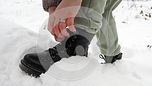 A woman laces up black winter boots on a street in a snowy forest. Women\'s hands tie shoelaces, winter shoes.