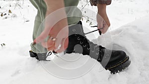 A woman laces up black winter boots on a street in a snowy forest. Women\'s hands tie shoelaces, winter shoes.