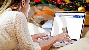 Young woman with laptop lying under Christmas tree and browsing obline store