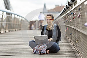 Woman with laptop sitting on a pedestrian bridge in an old European city.