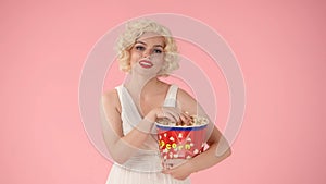 Woman with a large bucket of popcorn, enjoying watching a movie. Woman as Marilyn Monroe in studio on pink background.