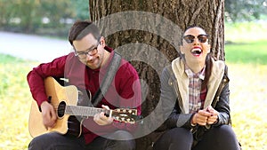 Woman laughing and man playing guitar while sitting on a tree in park