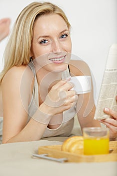 woman layed on bed eating breakfast and reading newspaper