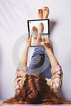 Woman lays with legs up on the wall and pushes buy now button on a laptop buying shoes. Online shopping concept.