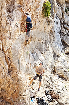 Woman lead climbing on natural cliff, guy belayer watching her