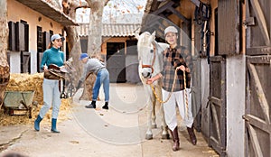 Woman leads a white horse by the bridle along stable