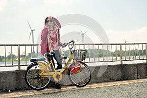 Woman lean with red coat to fence also hold hand of bicycle stand in front of wind turbines or windmill near water reservoir with