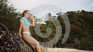 Woman leaning on a rock reef enjoying and relax warm dark golden sunset. Palm tree in beach. Woman in blue swimsuit and