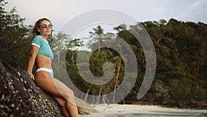 Woman leaning on a rock reef enjoying and relax warm dark golden sunset. Palm tree in beach. Woman in blue swimsuit and