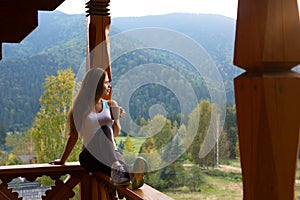 Woman leaning on wooden handrail and enjoys and relaxes beautiful mountain scenic. Young female on terrace sitting on