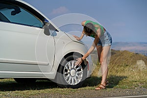 Woman leans down putting hand on wheel tire to check condition of white car parked on roadside