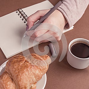 Woman left hand with pen writing on notebook at coffee shop. Freelancer working in outdoor. Coffe break with croissant and