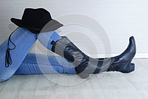 Woman legs in black cowboy boots with cowboy hat close-up. Sitting on gray wooden background